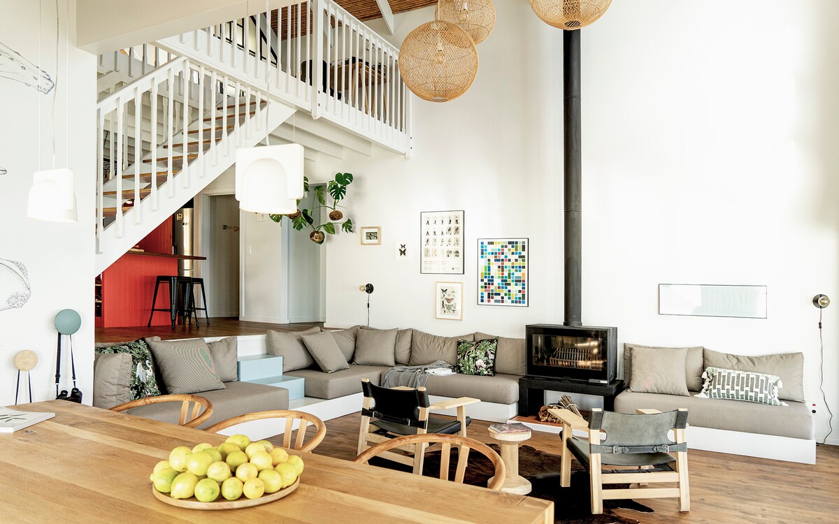 A spacious lounge area showcases a modern design with comfortable grey seating arranged around a black fireplace. Natural light fills the room, highlighting a large wooden dining table adorned with a bowl of lemons. A wooden staircase leads to the upper floor, adding charm to the space.