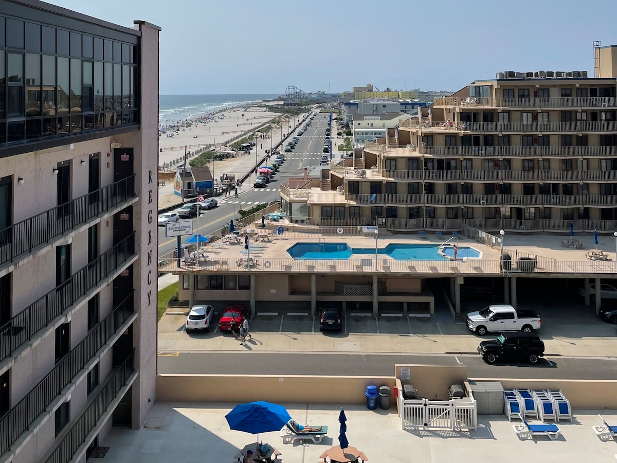 An aerial view showcases the Regency building with a rooftop pool deck. Below, several lounge chairs surround the pool area, and vehicles are parked in the lot. The beach is visible in the distance, with a boardwalk stretching along the shoreline.