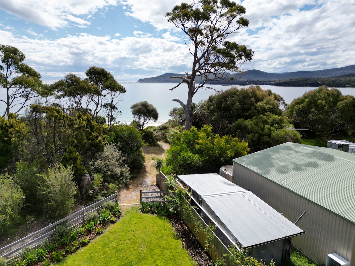 A breathtaking view of the serene bay, framed by lush greenery and towering trees. The coastline meets the calm water, with sandy shores visible in the distance. A well-maintained garden and structures are incorporated into the tranquil landscape, creating a peaceful atmosphere.