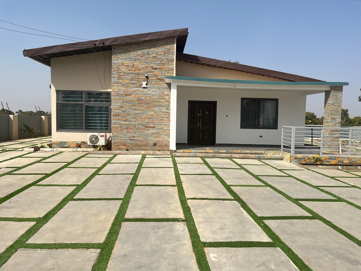 A modern single-story house features a stone façade and a sloped roof. A spacious front porch is complemented by a well-maintained stone pathway, adorned with green grass sections. Large windows allow natural light into the interior, enhancing the inviting appearance of the home.