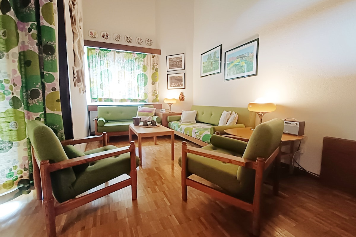 A welcoming living area is furnished with two green sofas and two matching armchairs. Wooden accents provide a warm touch. Natural light filters through a window adorned with patterned curtains, highlighting the decor, which includes framed artwork and decorative plates on the wall.