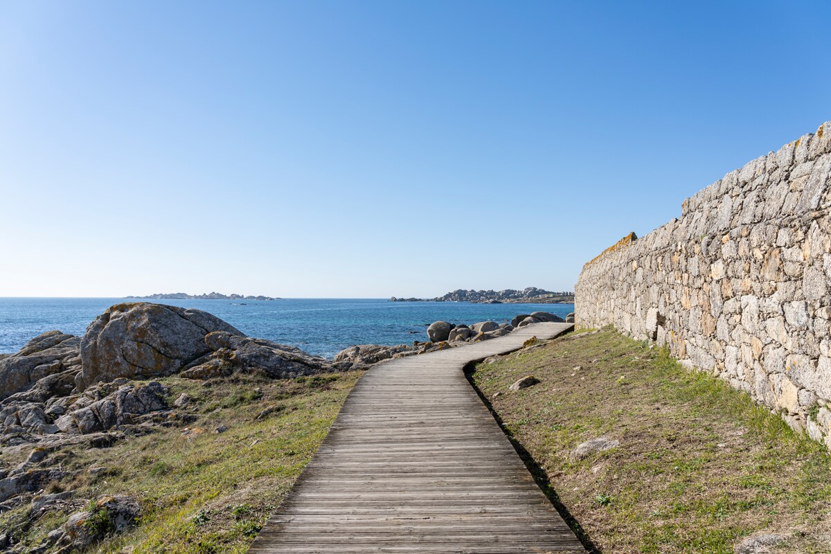 A wooden pathway winds along the coastline, bordered by a stone wall and stretching towards the horizon. On either side, rocky formations with patches of grass are visible, while the serene blue sea and clear sky create a tranquil backdrop.