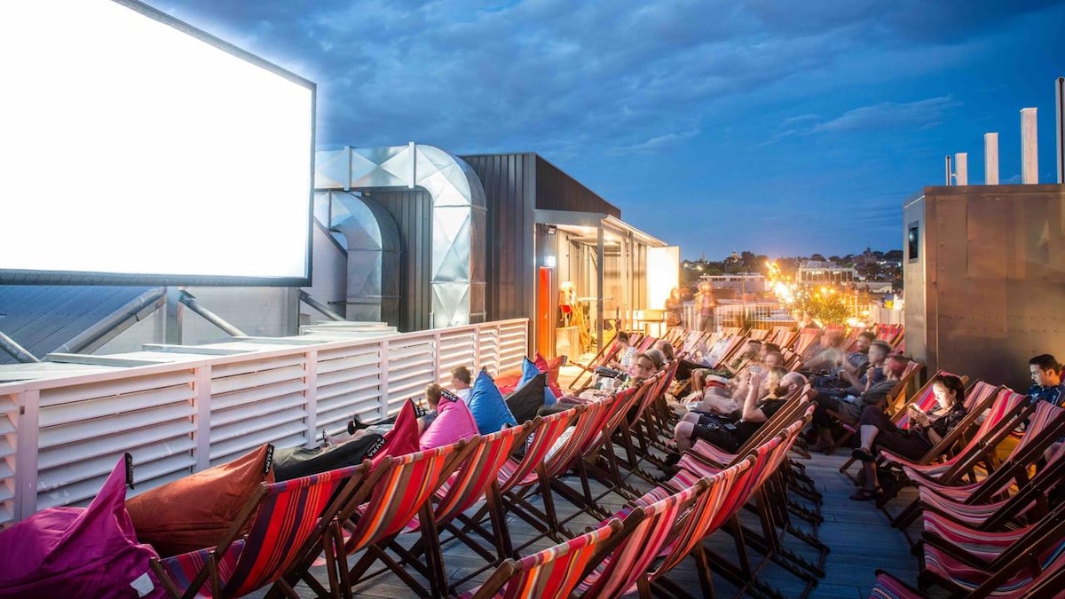A rooftop area features rows of striped lounge chairs, creating a relaxed setting for outdoor movie viewing. Soft bean bags in various colors are scattered throughout. A large screen is positioned at the front, with city lights twinkling in the background against a dusky sky.