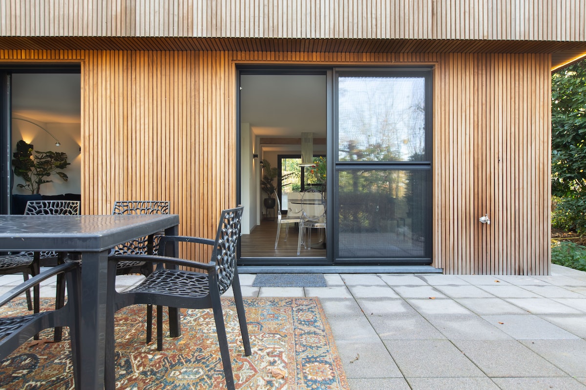 A patio with a dark wooden table and intricately designed black chairs is visible. Natural light enters through a large glass door, leading to a cozy interior. A patterned rug adds a touch of comfort on the stone floor, surrounded by greenery.