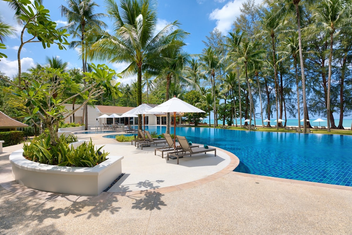 A tranquil outdoor swimming pool is surrounded by palm trees and tropical landscaping. Several lounge chairs with white umbrellas provide shaded seating. The clear water of the pool reflects the blue sky, creating a serene atmosphere. A beach area is visible in the background.