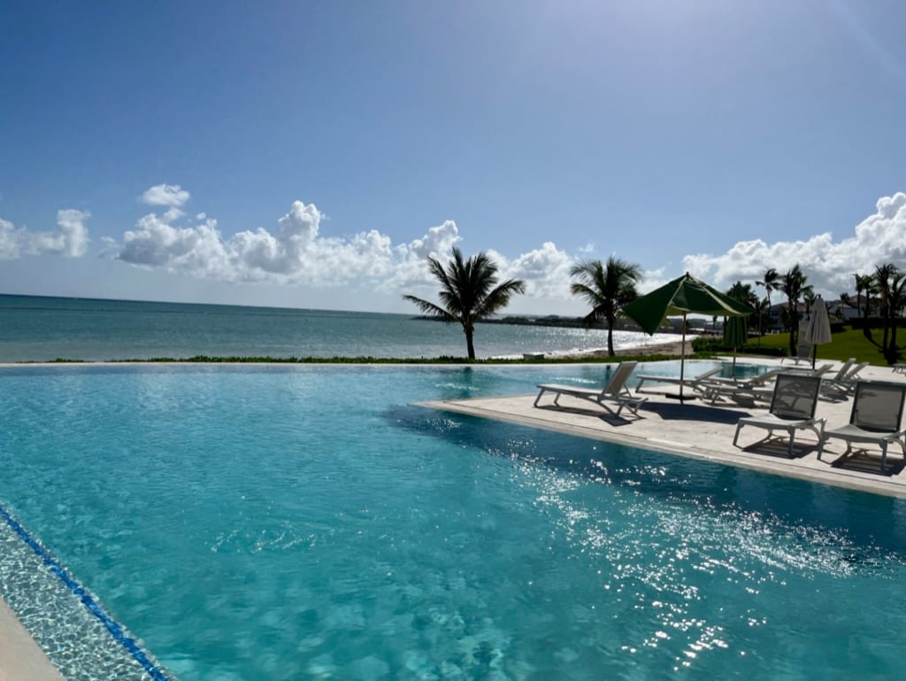 An inviting infinity pool is featured in the foreground, reflecting the clear turquoise water. Lush palm trees are visible in the distance, framed by a serene ocean and softly clouded sky.