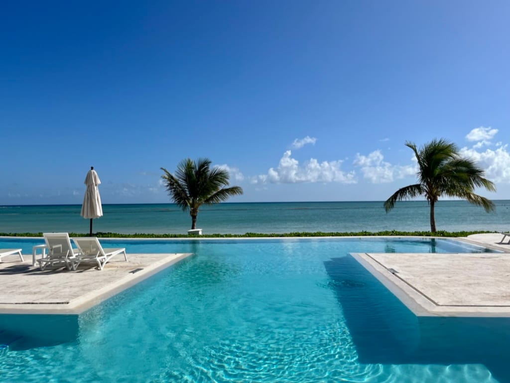 An inviting infinity pool extends towards the horizon, offering views of the turquoise sea. Two palm trees frame the scene, while a sun umbrella provides shade over a nearby lounge area. The clear blue sky enhances the tranquil ambiance of the beachfront setting.