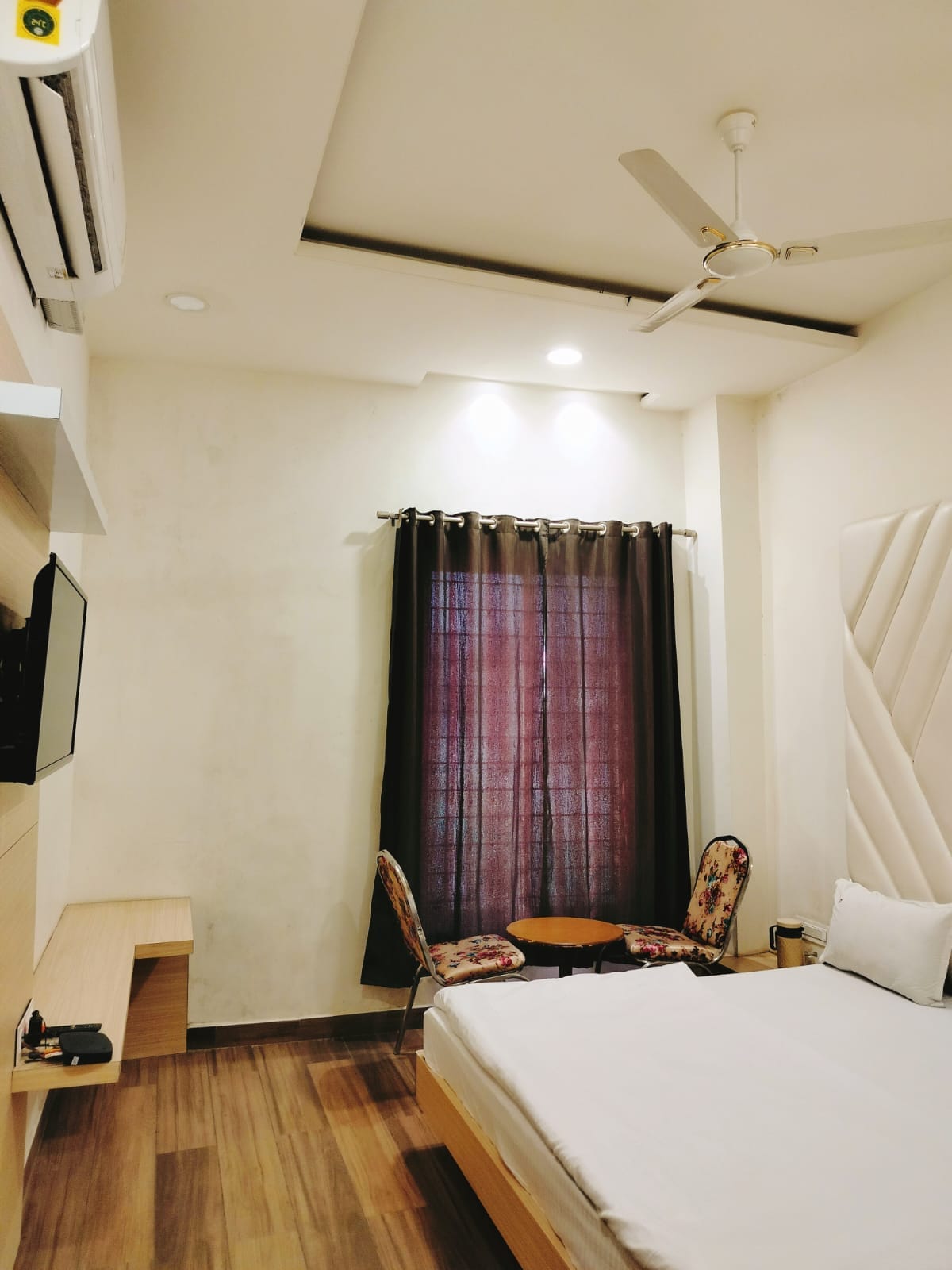 A spacious room features a king-sized bed covered with crisp linens. A small table with two chairs is positioned near a window, adorned with dark curtains. A wall-mounted television and a ceiling fan are present, contributing to a comfortable atmosphere.
