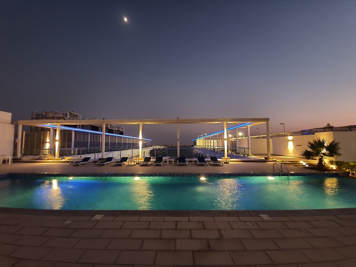 A rooftop swimming pool is illuminated under the evening sky, showcasing clear water reflecting soft lighting. Lounge chairs are positioned around the pool, providing spaces for relaxation. The backdrop includes a spacious deck area, enhancing the serene ambiance of the setting.