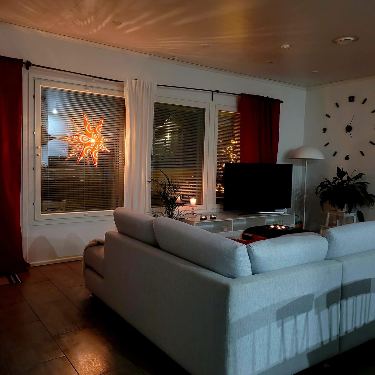 The living area displays a light gray sofa facing a television, with decorative elements visible such as a unique star-shaped lamp and plants. Large windows, adorned with white curtains, allow for ambient lighting, while warm-toned curtains provide contrast against the light walls.