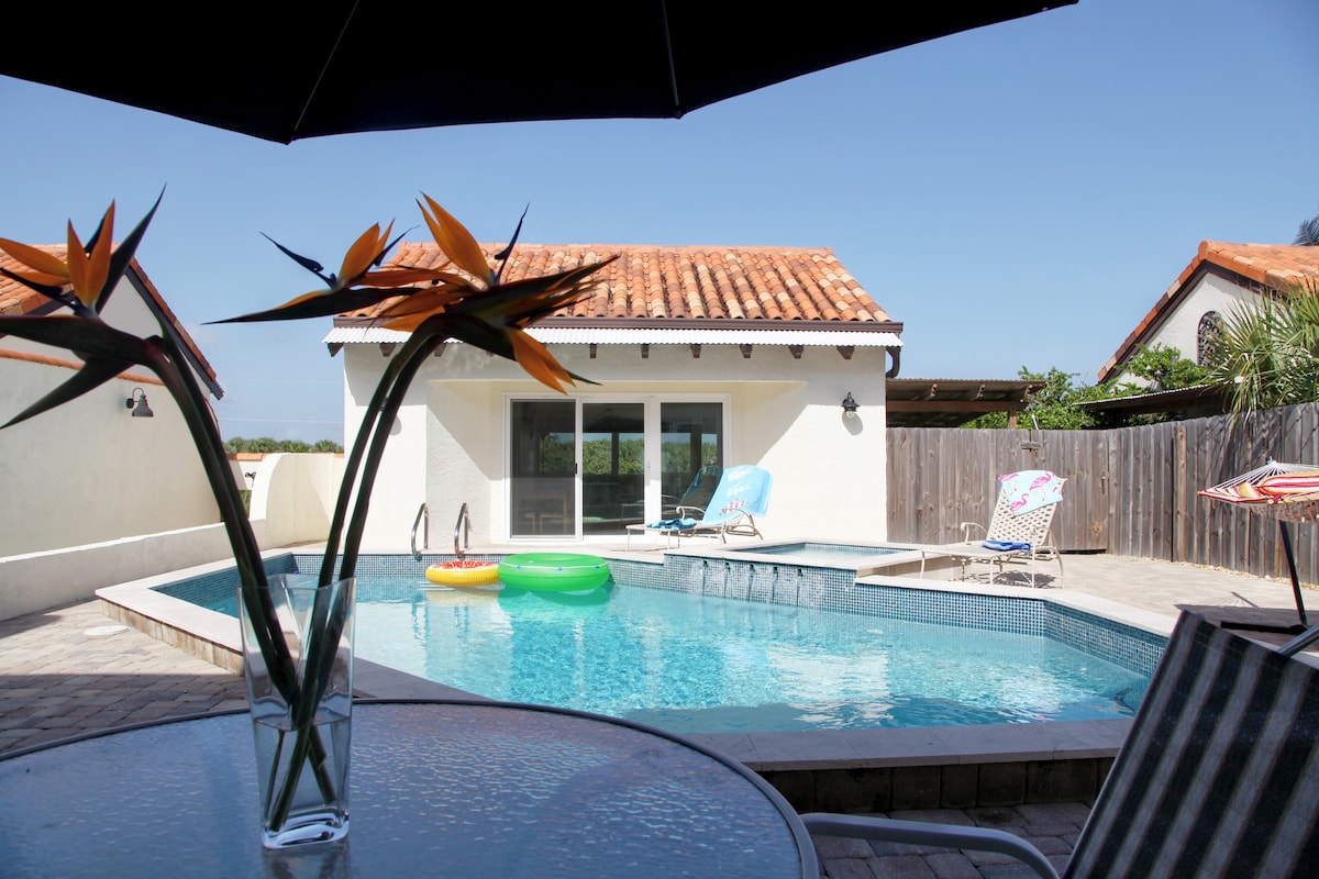 The outdoor space features a private pool surrounded by a patio area. A line of lounge chairs sits beside the water, while a large umbrella provides shade. In the foreground, orange bird-of-paradise flowers are displayed in a vase, enhancing the serene environment.