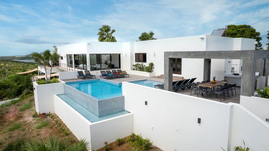 The villa is highlighted with an infinity pool that flows over a sleek edge, surrounded by lounge chairs for relaxation. A spacious terrace provides seating for outdoor dining. Large windows invite natural light, connecting the indoor and outdoor areas with scenic views of the surrounding landscape.