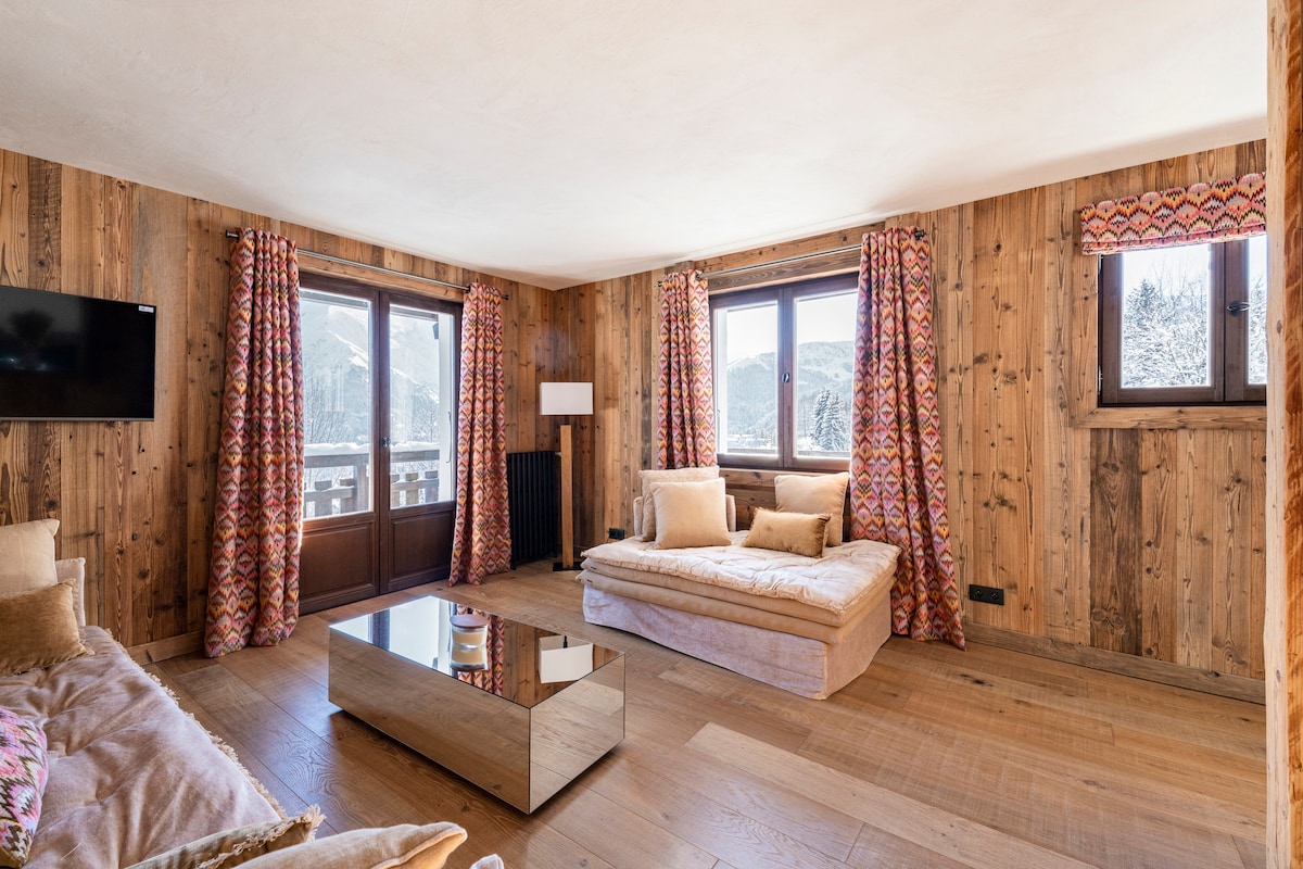 A living area features wooden walls and large windows that provide mountain views. Soft seating is arranged around a reflective coffee table. Colorful patterned drapes frame the windows, creating a cozy atmosphere with natural light enhancing the space.
