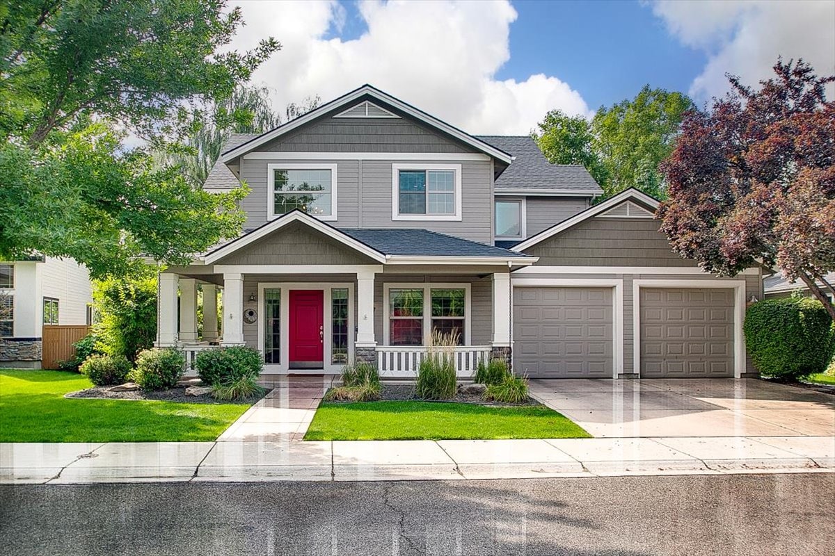 A two-story home is presented with a light gray exterior and a striking red front door. The covered porch frames the entrance, accompanied by a landscaped area featuring various shrubs and trees. A double garage is available, complementing the home's welcoming appearance.