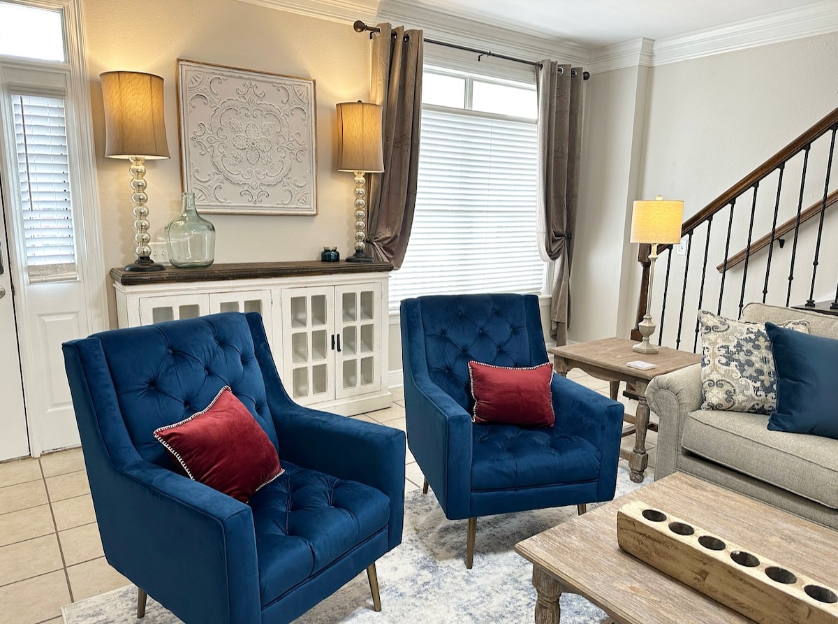 Two blue armchairs are positioned in a welcoming living room, accented by red throw pillows. A wooden coffee table sits in front of a light-colored sofa, with decorative items displayed on a console table against the wall. Natural light filters through the window, enriching the space.