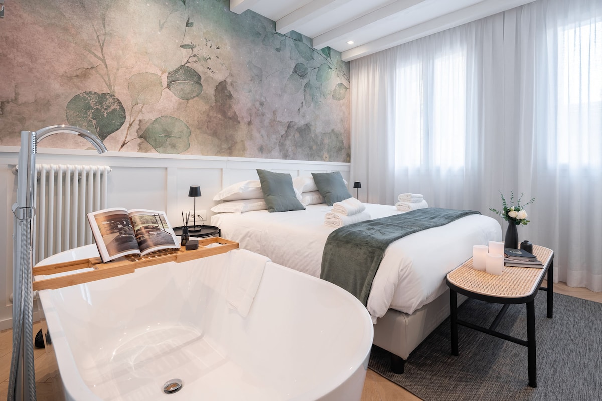 A spacious bedroom is highlighted by a soft color palette and large windows allowing natural light. A comfortable bed is accompanied by decorative cushions and a throw blanket. A standalone bathtub is positioned nearby, with a stylish table and an open book, enhancing a serene ambiance.