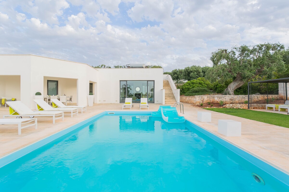 An outdoor area features a sparkling blue swimming pool, complemented by lounge chairs positioned around it. An inviting water slide is visible, with grassy space and trees in the background. The modern structure showcases large glass doors leading to the interior.