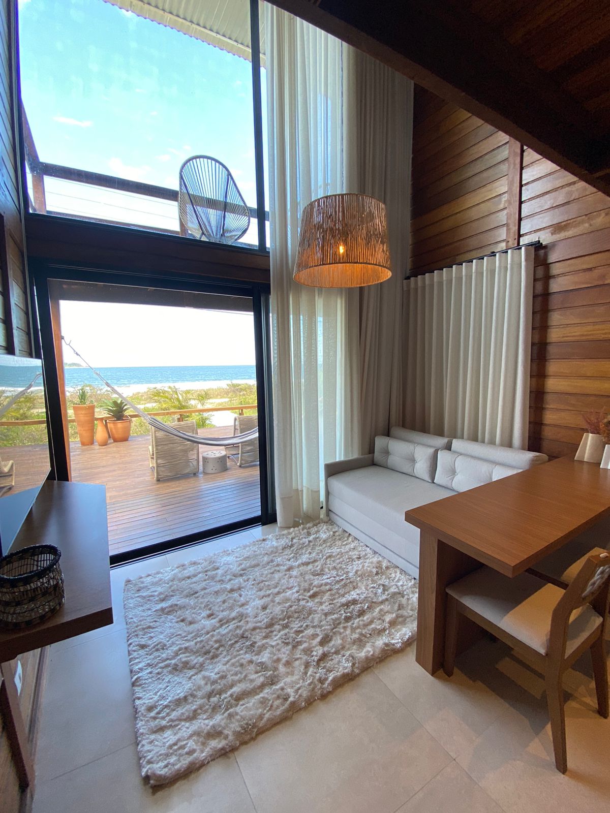 A bright and airy living area features large windows that offer a view of the sea. A comfortable white sofa and wooden dining table are complemented by a soft area rug. Natural light fills the space, enhancing the wooden wall accents.