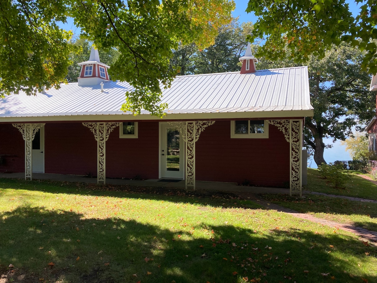 The exterior of Willow Lodge North is showcased with a charming red facade and decorative white trim. Features include two distinctive cupolas atop a metal roof, flanked by large windows that invite natural light. Lush greenery surrounds the building, providing an inviting view of the landscape.