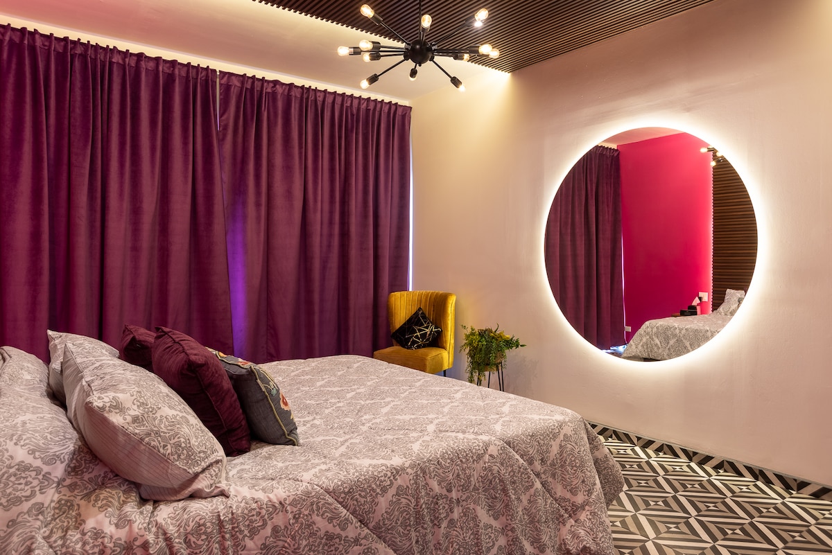 A spacious bedroom is presented with a large bed draped in patterned linens. Rich purple curtains frame the window, complementing a bright pink accent wall. A round mirror illuminated by soft light hangs prominently. A yellow armchair and a plant add subtle accents to the room.