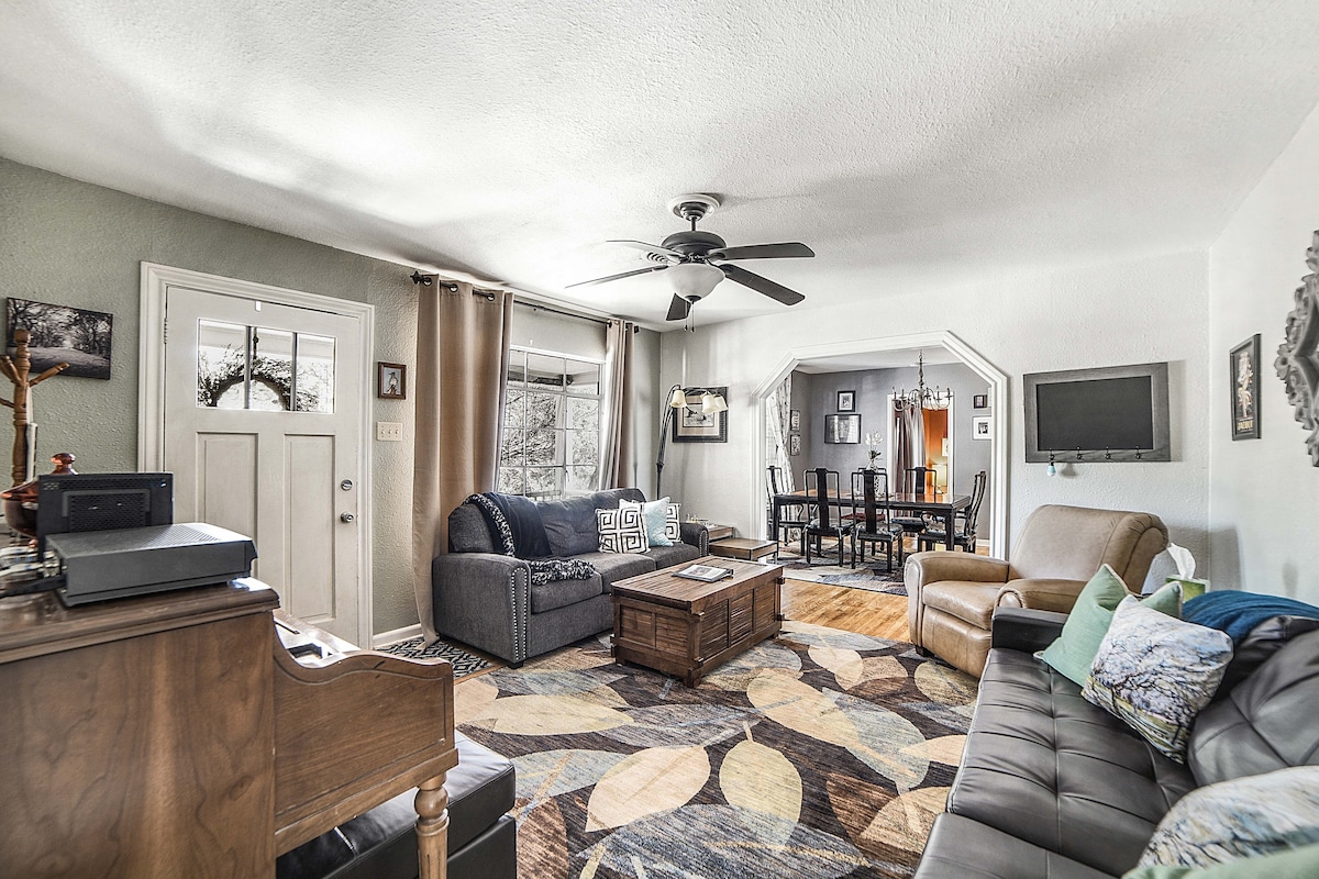 A living area is shown, featuring a ceiling fan, two sofas, and a coffee table surrounded by a floral-patterned rug. Natural light enters through the front door and windows, with artwork adorning the walls. A dining area can be seen in the background.