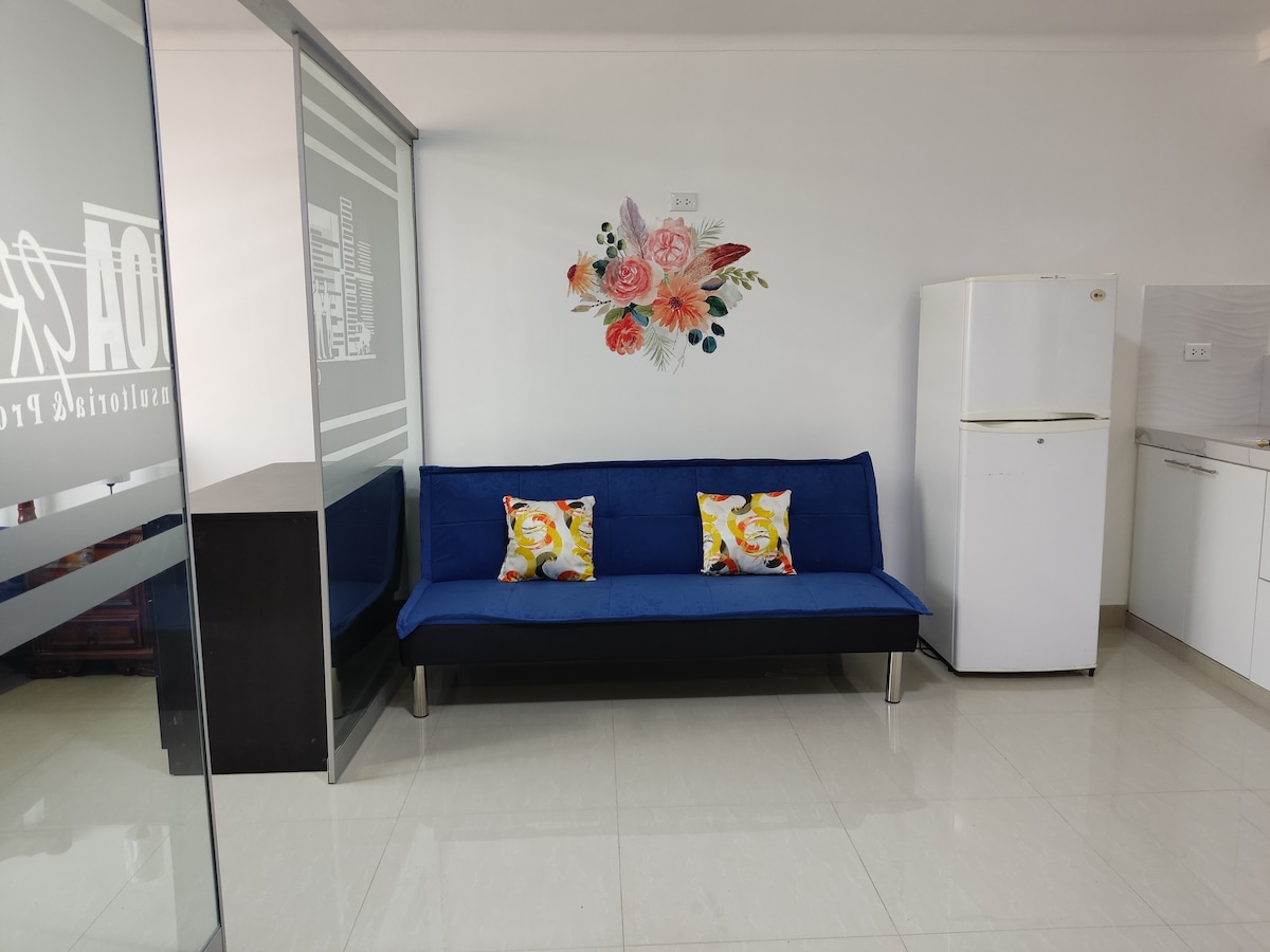 A cozy seating area is presented with a blue sofa adorned with colorful floral cushions. A decorative wall art piece featuring flowers adds character. A white refrigerator stands nearby, contributing to the functional kitchen space, while glossy tiles reflect natural light.