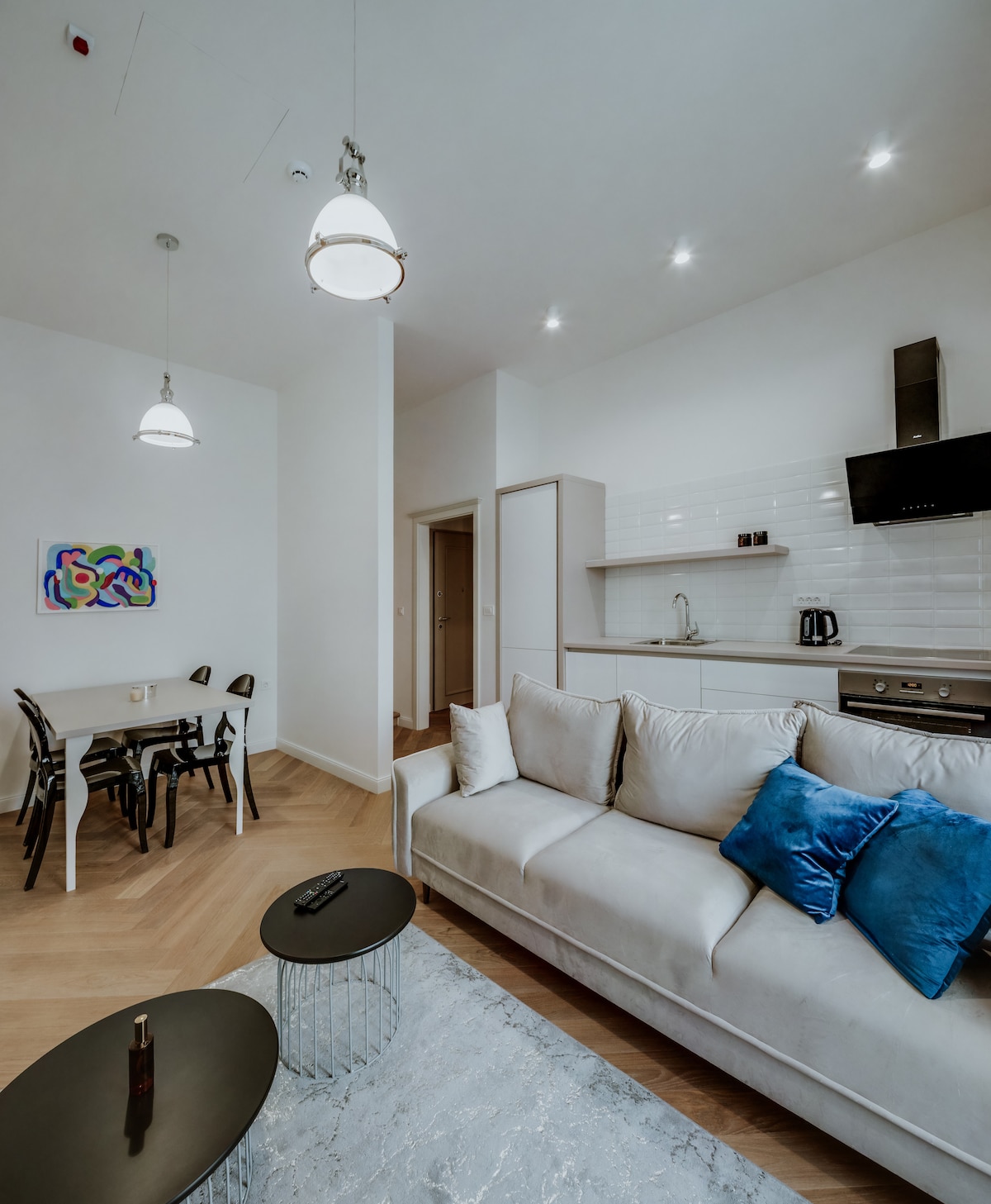A modern living area features a beige sofa accented with blue pillows, accompanied by two round glass tables. A dining table with black chairs is situated nearby, with a kitchen area visible in the background, showcasing clean lines and minimalist appliances.
