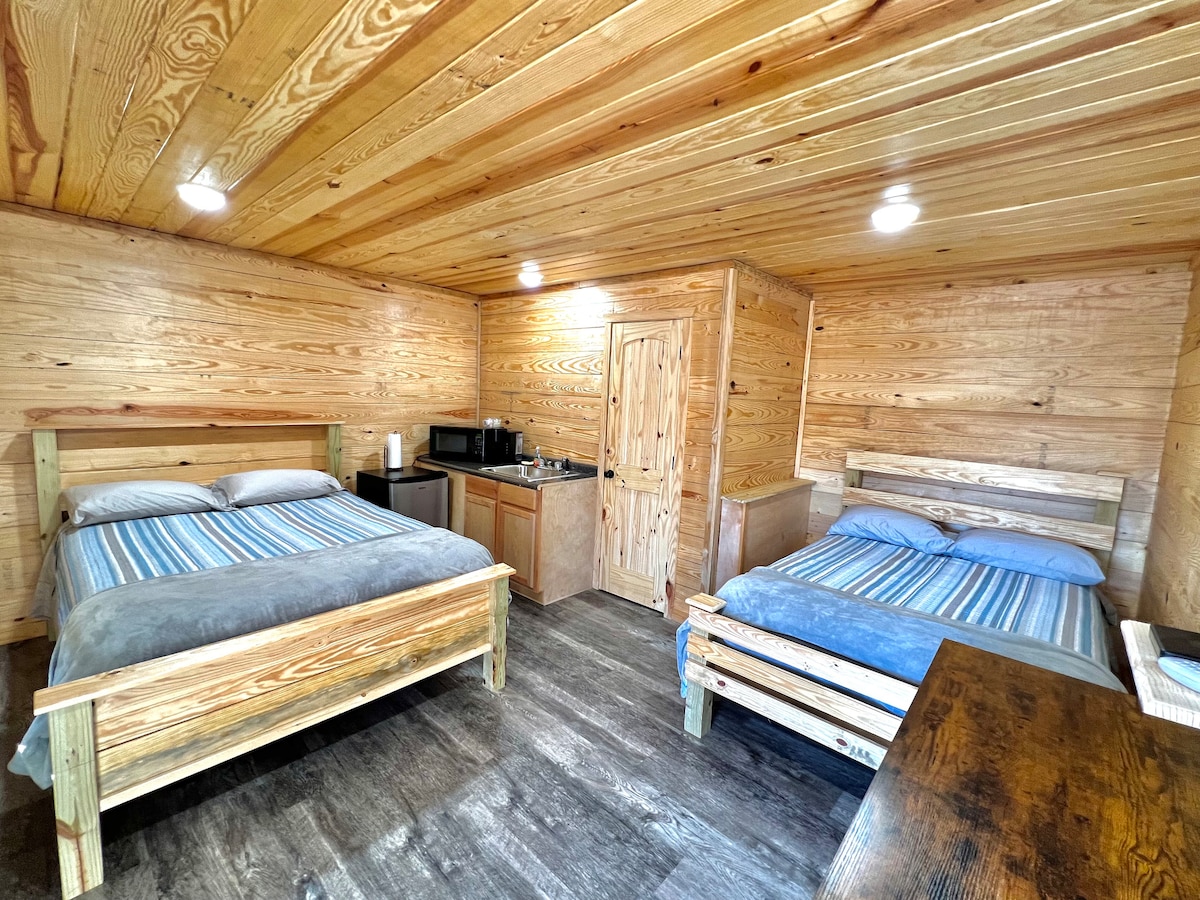A cabin room features two beds with blue and gray striped linens, complemented by a wooden table and a kitchenette with modern appliances. Light is provided by recessed ceiling fixtures, illuminating the natural wood paneling throughout the space. A door leads to additional facilities.