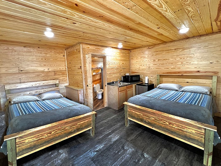A spacious cabin interior features two beds with neutral bedding and striped accents. Wooden walls contribute to a rustic ambiance, while a kitchenette is visible in the background with modern appliances. A bathroom door is positioned at the rear, providing convenient access.