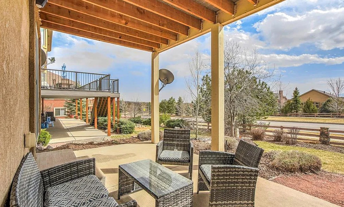 An outdoor patio area features comfortable seating with woven chairs and a glass table. The space is shaded by a wooden roof and overlooks well-maintained landscaping. Nearby, a pathway leads to the entrance, with open views of the surrounding neighborhood.