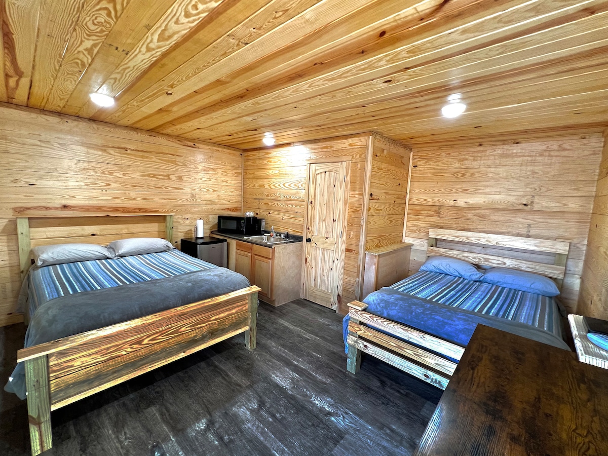 The interior of the cabin features two spacious beds with striped bedding and wooden frames. A kitchenette area is visible, equipped with a microwave and a small refrigerator. Wood-paneled walls and ceiling create a warm atmosphere, complemented by soft lighting from overhead fixtures.