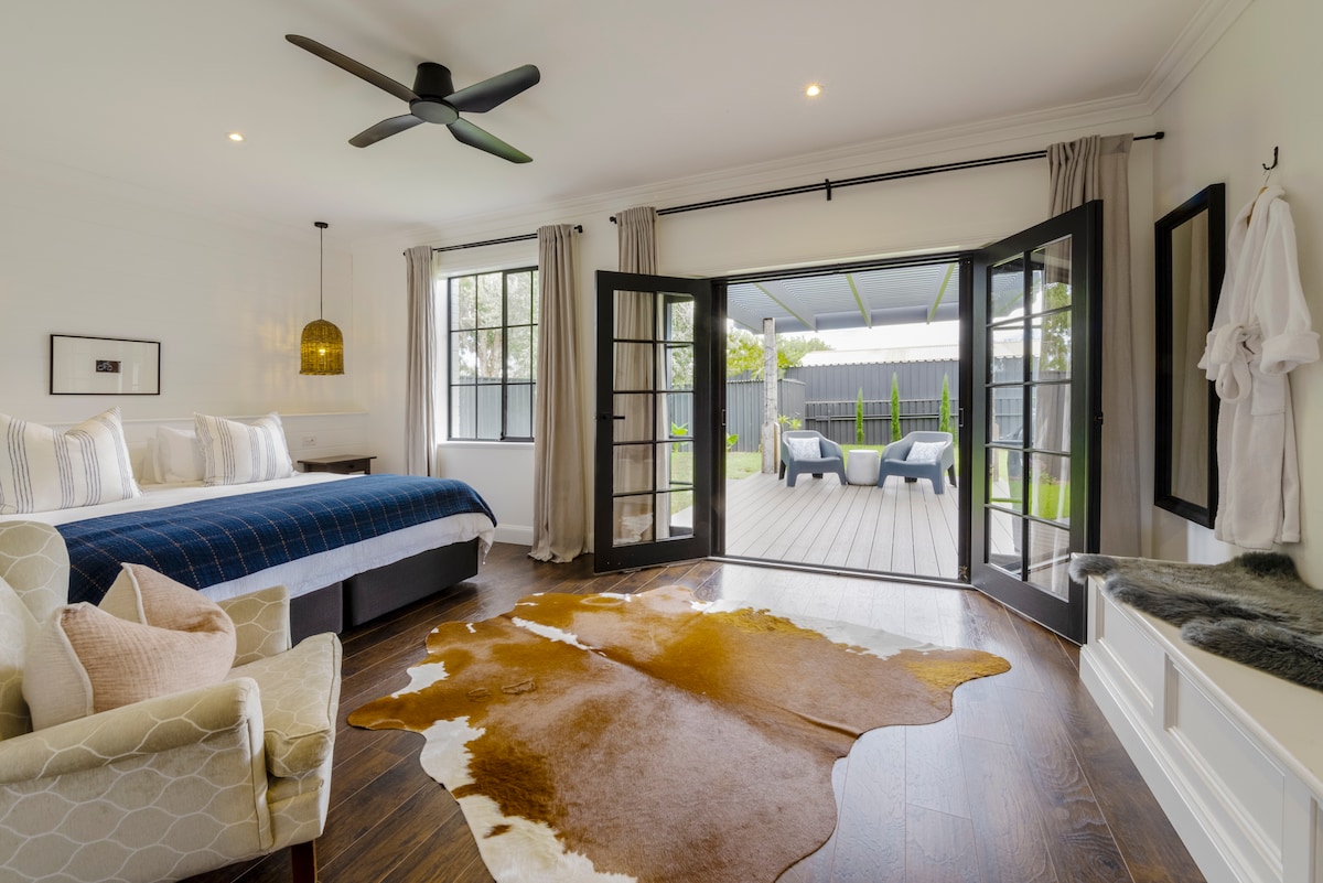 The king bedroom features large glass doors that open to a spacious deck. A cozy seating area includes a light-colored armchair, and a patterned rug adds texture to the wooden floor. Natural light illuminates the room, emphasizing the neutral decor and inviting ambiance.