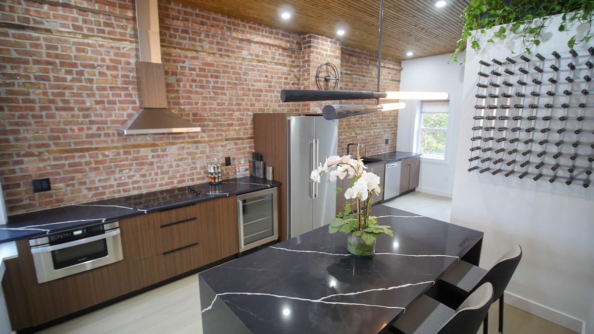 A modern kitchen features exposed brick walls, sleek cabinetry, and a large dark countertop with a striking white veining. Stainless steel appliances are incorporated, and a floral arrangement adds a touch of greenery. Ample natural light filters through the nearby window.