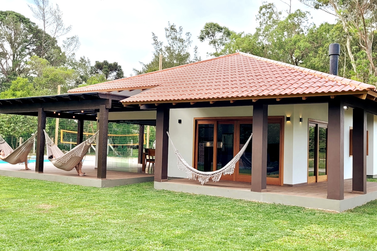 A modern house features a spacious exterior with a tiled roof, set amidst lush greenery. Hammocks are suspended on the front porch, inviting relaxation. The area includes a view of the swimming pool and volleyball court, creating a welcoming outdoor space.