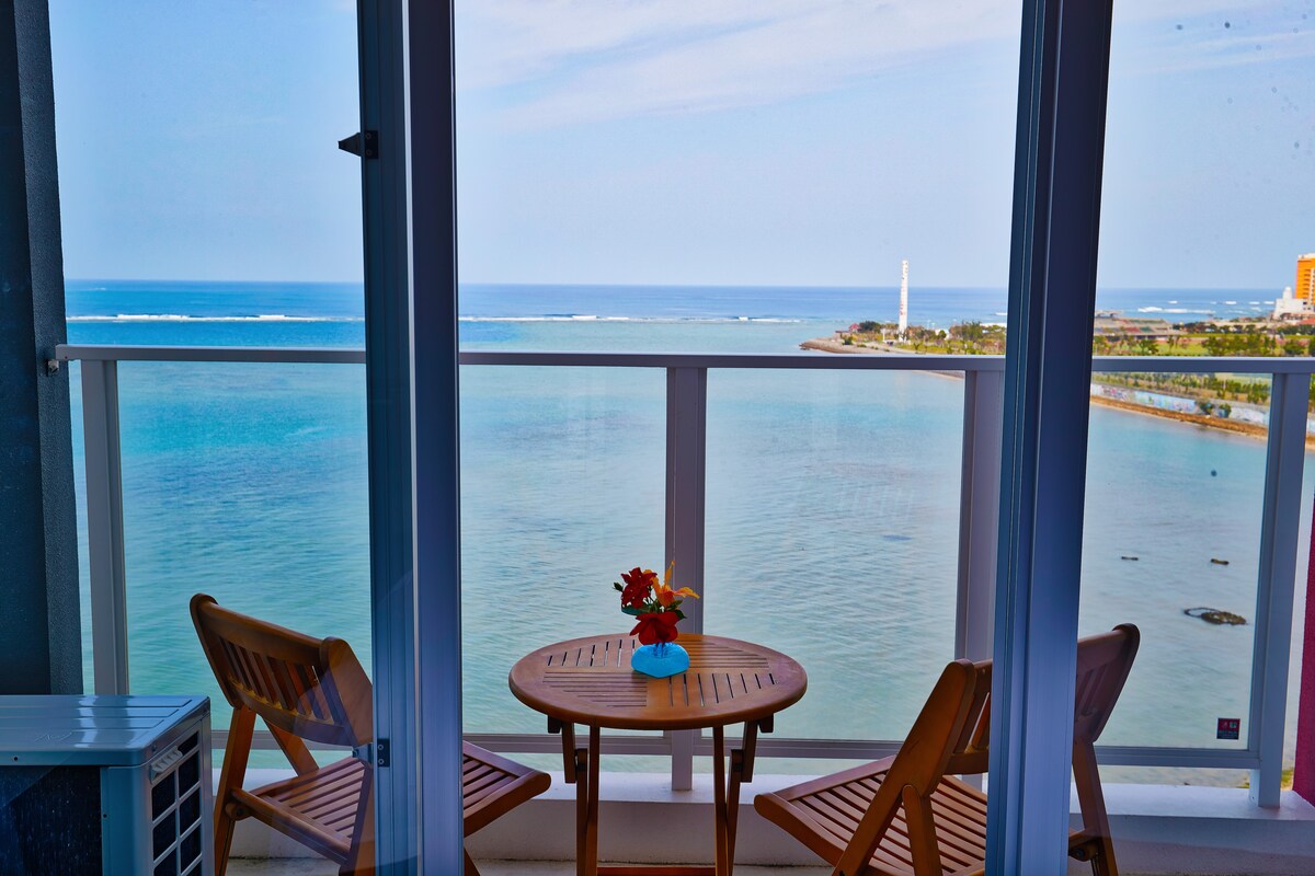 A private balcony features a wooden table and two matching chairs, offering an unobstructed view of the ocean. A small bouquet of flowers sits at the center of the table. The coastline and horizon are visible in the distance under a clear blue sky.