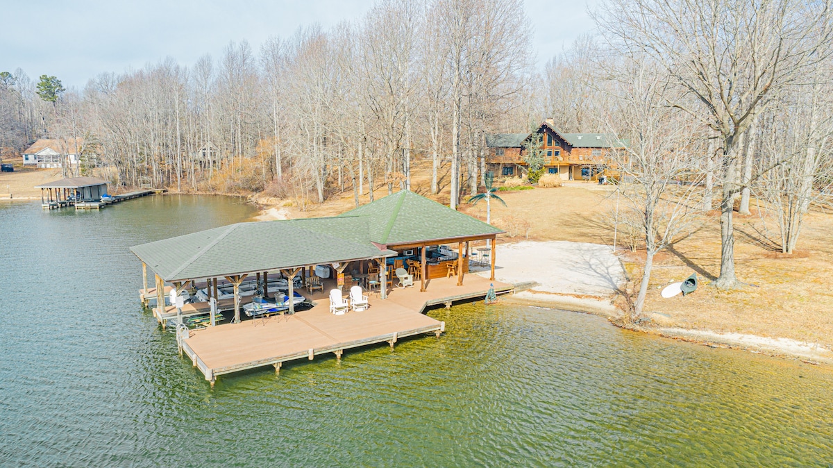A spacious private dock extends over the water, featuring a covered area with seating for relaxation. Surrounded by trees, the property is nestled by the lakeside, offering clear views of the log cabin home set in the background against a serene natural landscape.