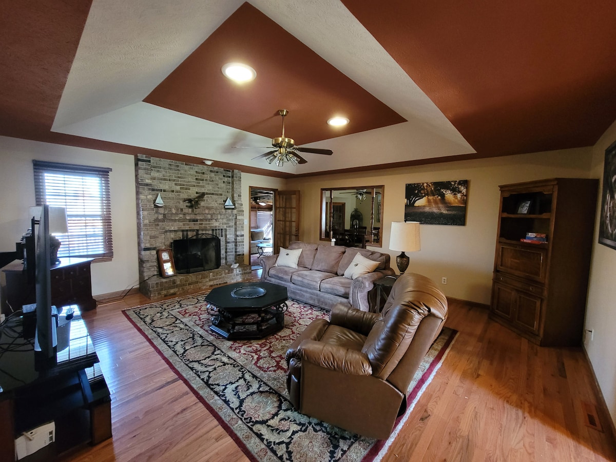 A spacious living area is highlighted by warm earth tones and natural light. A comfortable sofa faces a large flat-screen TV, with a patterned area rug anchoring the conversation space. A brick fireplace adds texture, and bookshelves flank the room, providing ample storage.