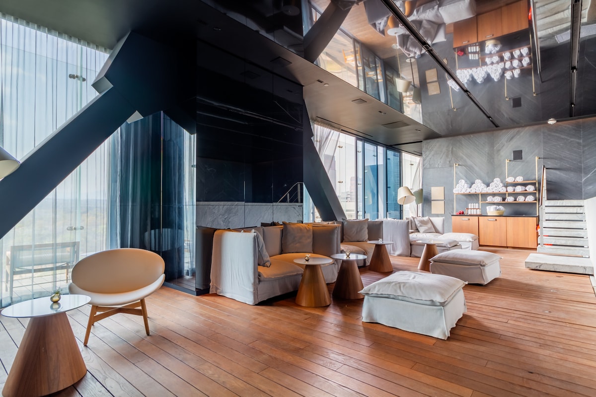 A spacious lounge area features comfortable seating arrangements with light-colored cushions, complemented by wooden accent tables. Large windows create an open feel, allowing natural light to illuminate the space, while elegant decor elements enhance the modern design.