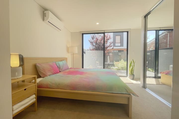 A bright bedroom with a wooden bed frame and colorful tie-dye bedding. The room features large sliding glass doors that open to a patio area with outdoor seating. A potted plant sits in the corner, and an air conditioning unit is mounted on the wall.