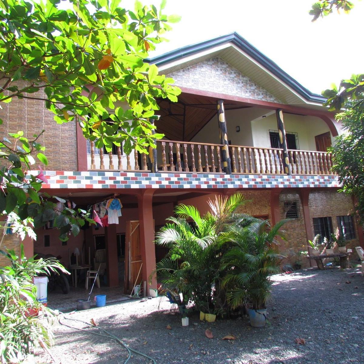A two-story building is surrounded by lush greenery, featuring a balcony with decorative railings. The exterior highlights a mix of brick and vibrant tile patterns. Hanging clothes can be seen, adding a lived-in feel to the outdoor space.