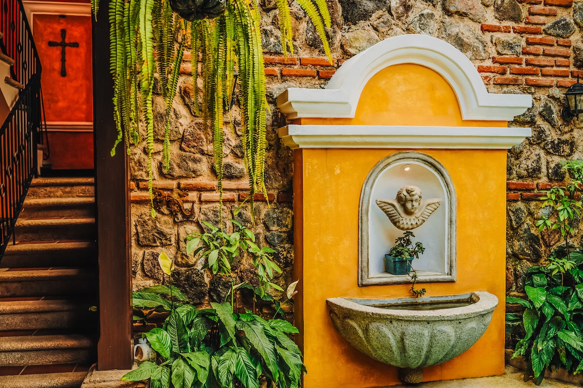 A vibrant orange fountain is featured, framed by stone walls and lush greenery. A bird sculpture is positioned at the center of the fountain, complemented by a small plant. Stairs lead to a door adorned with a cross in the background, enhancing the setting's character.