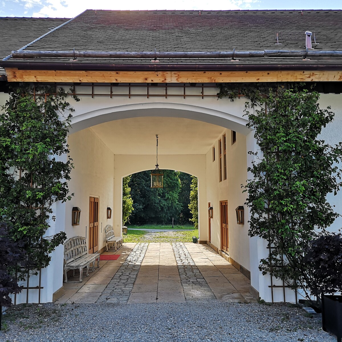 An arched entryway is flanked by greenery, leading to an open courtyard. Stone paving is visible, lined with benches on either side. Lanterns hang from the ceiling, providing soft illumination. Lush trees can be seen through the arch, creating a serene backdrop.