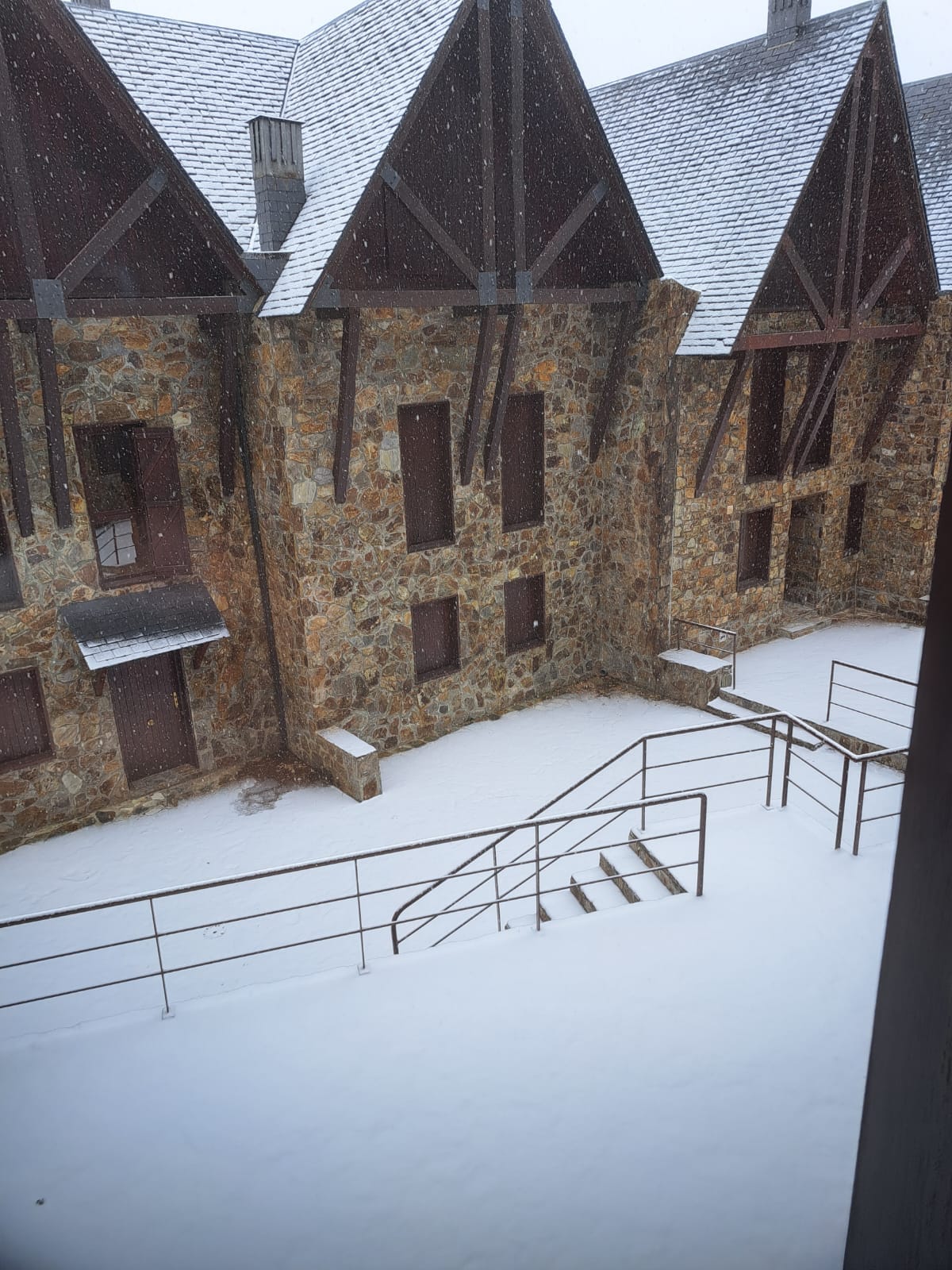 A snow-covered courtyard is visible through a window, with stone buildings featuring steep roofs in the background. Light snow is falling, creating a serene atmosphere. A set of metal stairs leads down from the foreground, surrounded by a layer of fresh snow.