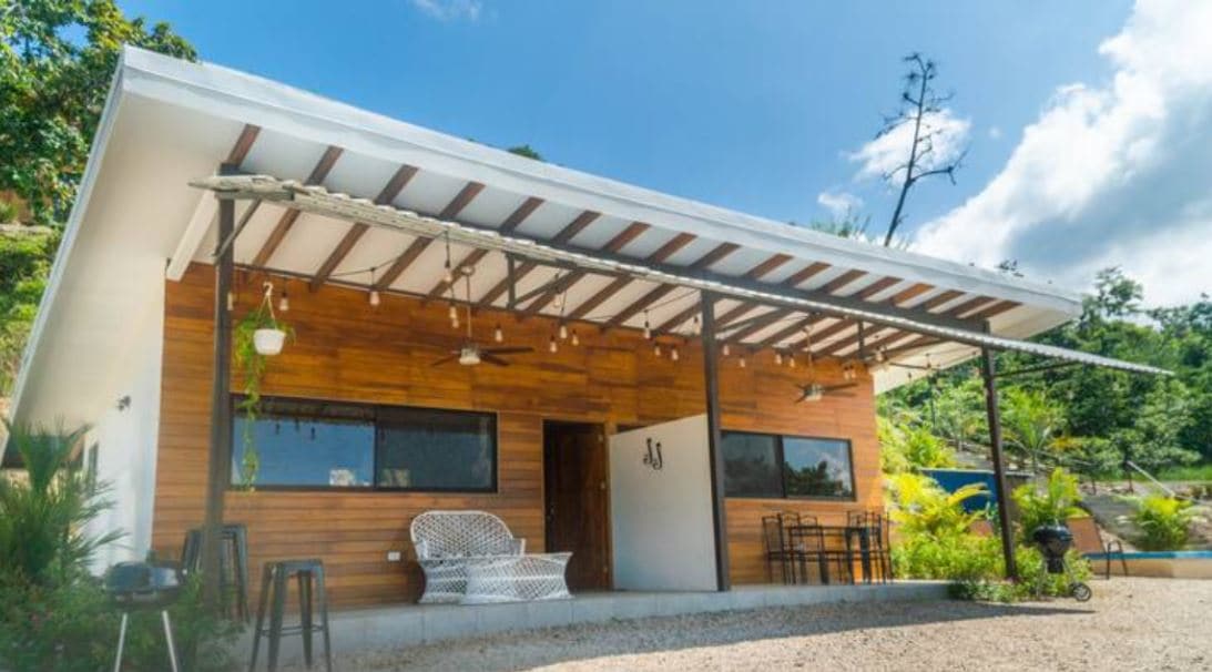A modern cabin features a wooden exterior with large windows providing natural light. An inviting covered porch is equipped with seating arrangements. Lush greenery surrounds the property, enhancing the connection with nature. The roofline adds a contemporary touch to the overall design.