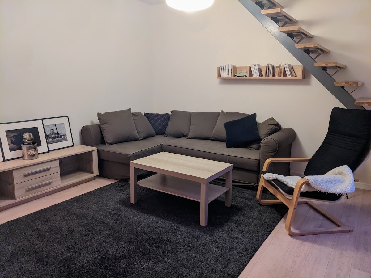 A cozy living area features a dark sofa complemented by decorative pillows, a light wood coffee table, and a low cabinet displaying framed photographs. A plush dark rug anchors the space, while a staircase ascends to a second level, illuminated by a round ceiling light.