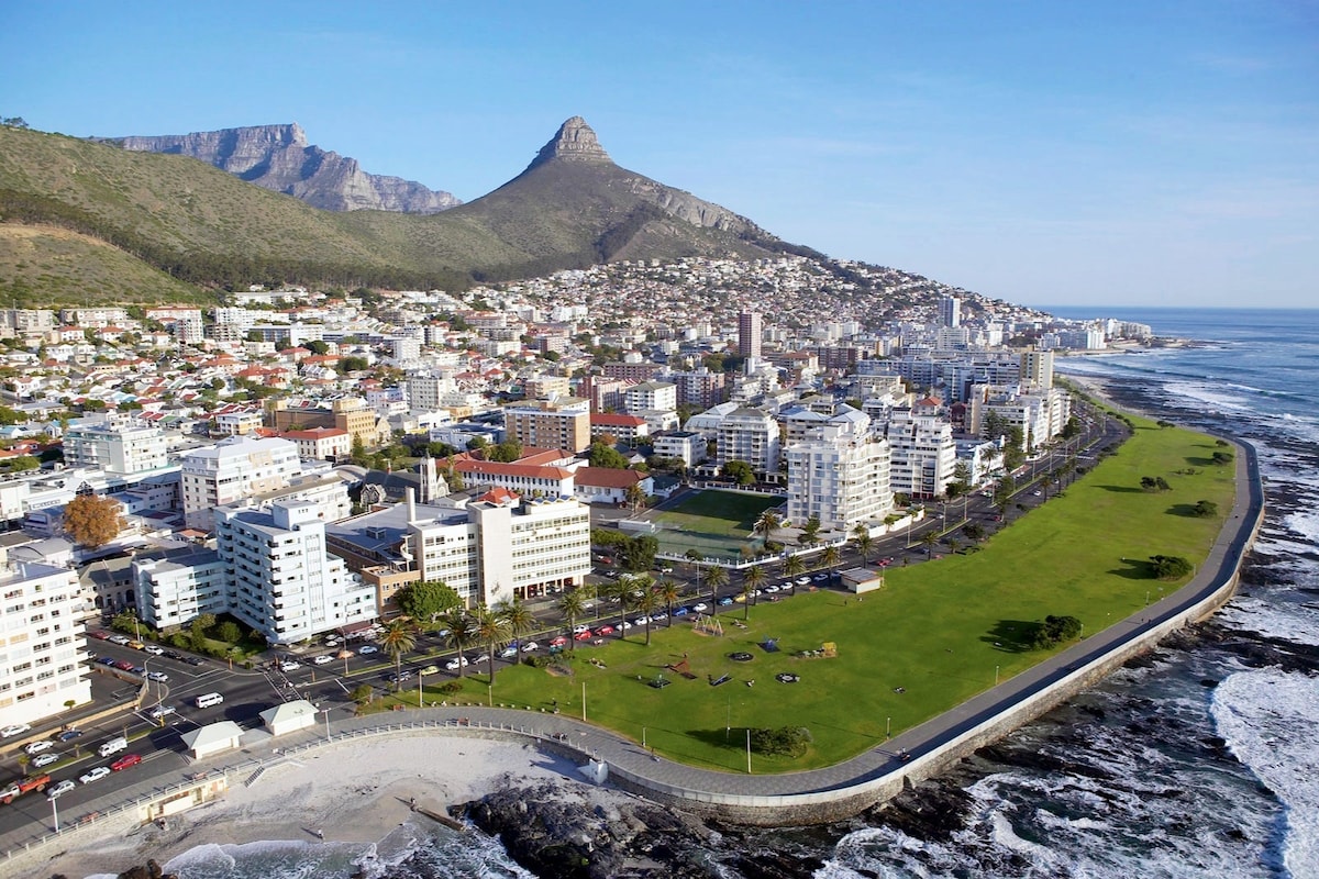 Aerial view of a coastal city, showcasing a vibrant promenade lined with green lawns and pedestrian pathways. The city's buildings are nestled against the backdrop of mountains, with the ocean visible on one side. Roads are seen with flowing traffic, and the scene is bathed in natural light.