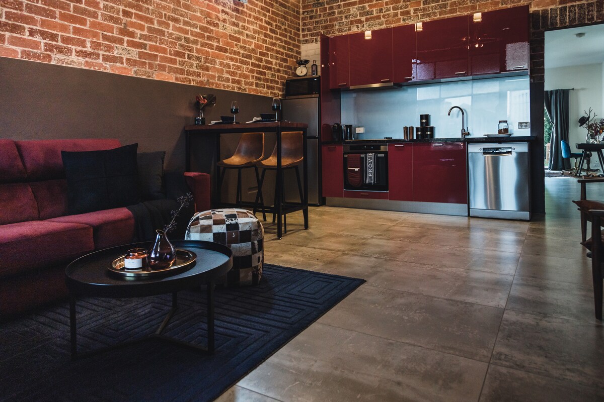An open kitchen and living area features exposed brick walls and modern cabinetry. A cozy seating arrangement is complemented by a round coffee table. Bar stools are positioned at a counter, and natural light is visible from an adjacent room.