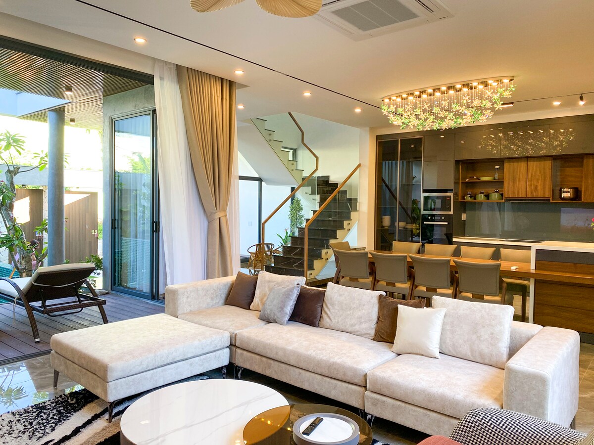 A modern living room features a spacious sectional sofa with neutral tones and decorative pillows. A stylish chandelier hangs overhead, illuminating the area. A dining table with matching chairs is positioned nearby, while large windows offer views of the outdoor space and natural light.