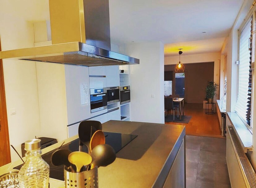 A modern kitchen is highlighted by sleek cabinetry and stainless-steel appliances. A central island features cooking utensils, while warm lighting illuminates the dining area in the distance, creating a welcoming ambiance.