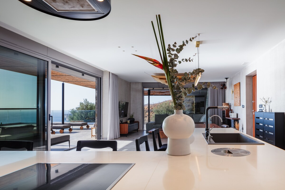 An open-plan living area is visible, featuring floor-to-ceiling glass doors that provide a seamless transition to an outdoor terrace. A modern kitchen island with a sleek design is complemented by a decorative vase holding greenery. Natural light fills the space, highlighting its minimalist style.
